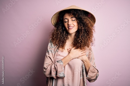 Young beautiful woman with curly hair and piercing wearing traditional asian conical hat smiling with hands on chest with closed eyes and grateful gesture on face. Health concept.
