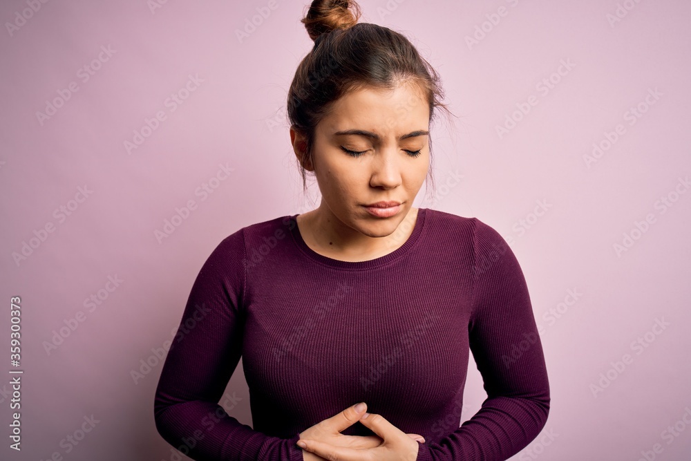 Obraz premium Beautiful young woman wearing casual bun hairstyle over pink isolated background with hand on stomach because indigestion, painful illness feeling unwell. Ache concept.