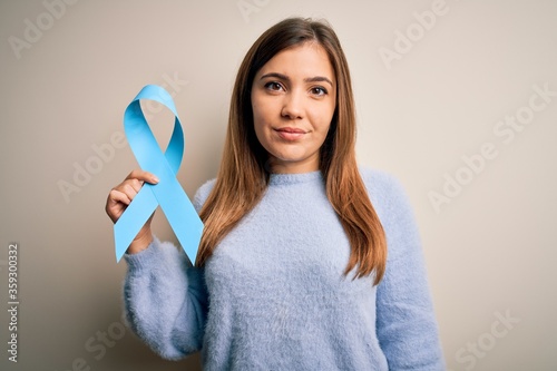 Young blonde woman holding prostate cancer awareness campaing blue ribbon with a confident expression on smart face thinking serious