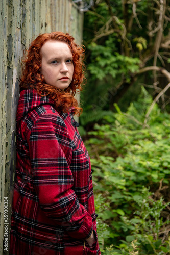 Portrait of a young woman with curly red hair, wearing a red tartan coat, set in a rough urban setting.