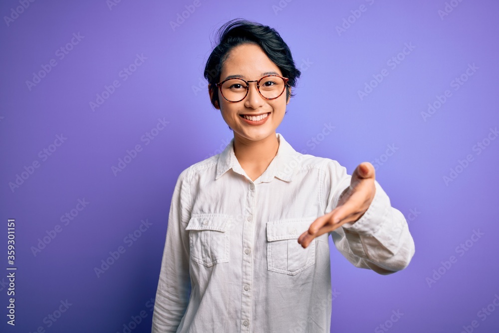 Young beautiful asian girl wearing casual shirt and glasses standing over purple background smiling friendly offering handshake as greeting and welcoming. Successful business. Young beautiful asian girl wearing casual shirt and glasses standing over purple background smiling friendly offering handshake as greeting and welcoming. Successful business.