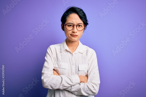 Young beautiful asian girl wearing casual shirt and glasses standing over purple background skeptic and nervous, disapproving expression on face with crossed arms. Negative person.