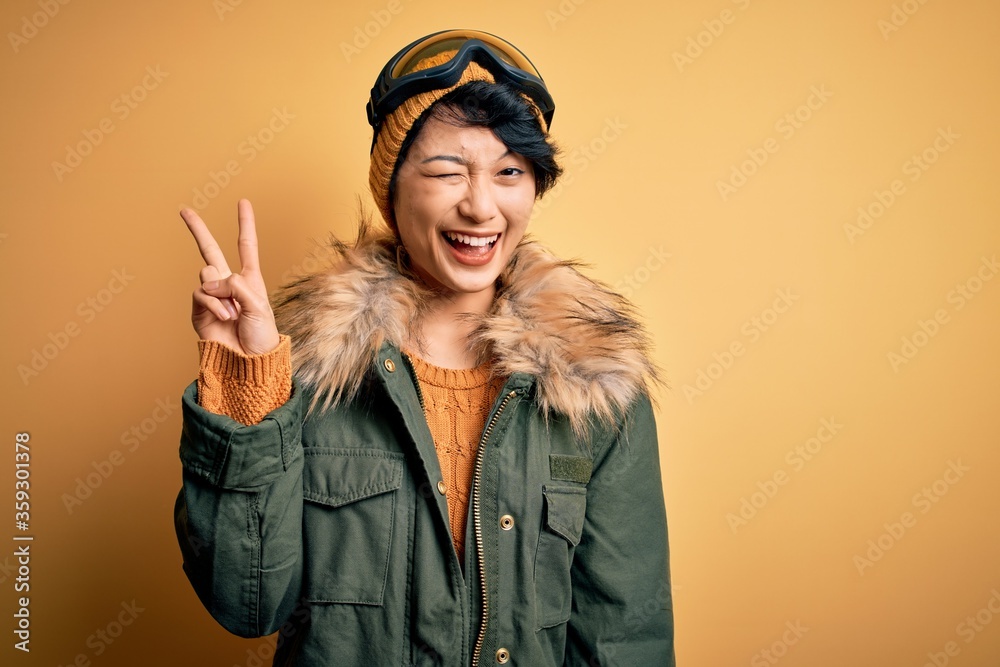 Beautiful asian skier girl wearing snow sportswear using ski goggles over yellow background smiling with happy face winking at the camera doing victory sign. Number two.