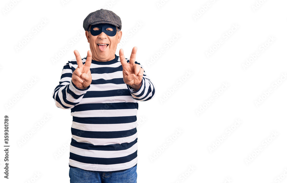 Senior handsome man wearing burglar mask and t-shirt smiling with tongue out showing fingers of both hands doing victory sign. number two. Senior handsome man wearing burglar mask and t-shirt smiling with tongue out showing fingers of both hands doing victory sign. number two.