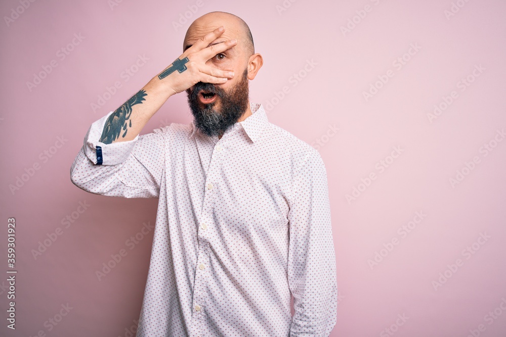 Handsome bald man with beard wearing elegant shirt over isolated pink background peeking in shock covering face and eyes with hand, looking through fingers with embarrassed expression. Handsome bald man with beard wearing elegant shirt over isolated pink background peeking in shock covering face and eyes with hand, looking through fingers with embarrassed expression.