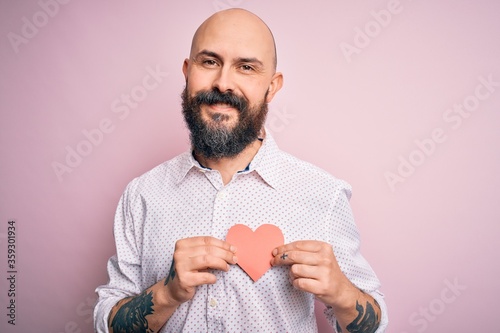 Handsome romantic bald man with beard holding red heart paper over pink background with a happy face standing and smiling with a confident smile showing teeth photo