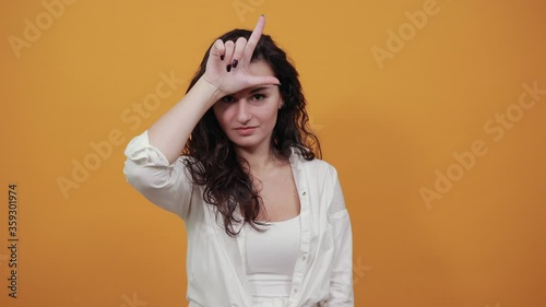 Showing loser sign, displaying dud gesture, alpha. Two fingers 2, disgust on face, negative human emotion facial expression. Young attractive woman, dressed white blouse, with brown eyes, curly hair