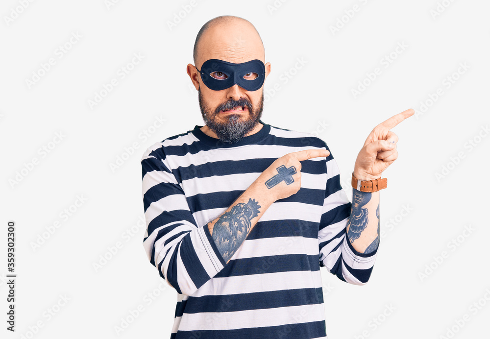 Naklejka premium Young handsome man wearing burglar mask pointing aside worried and nervous with both hands, concerned and surprised expression