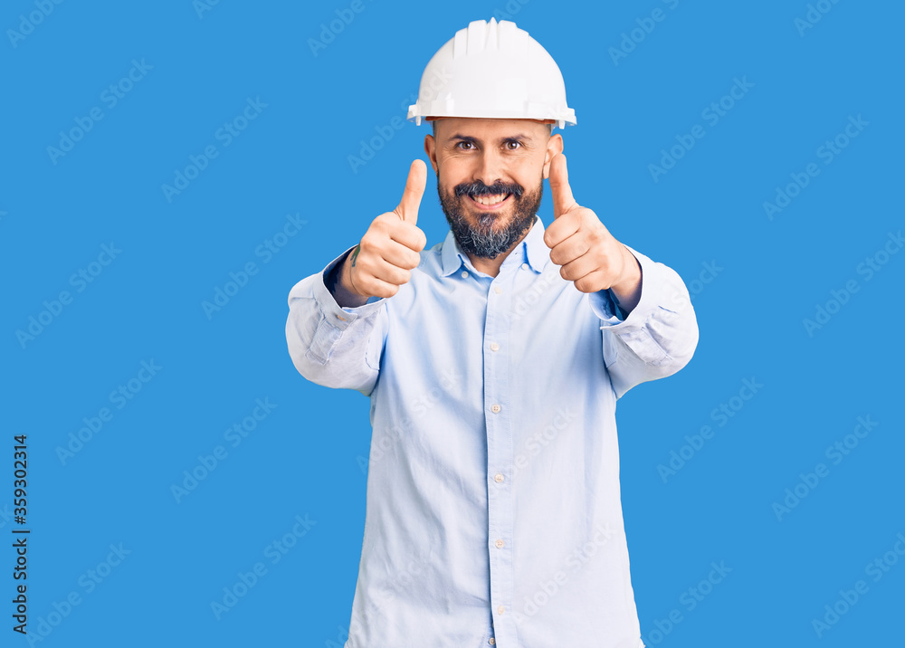 Young handsome man wearing architect hardhat approving doing positive gesture with hand, thumbs up smiling and happy for success. winner gesture. Young handsome man wearing architect hardhat approving doing positive gesture with hand, thumbs up smiling and happy for success. winner gesture.