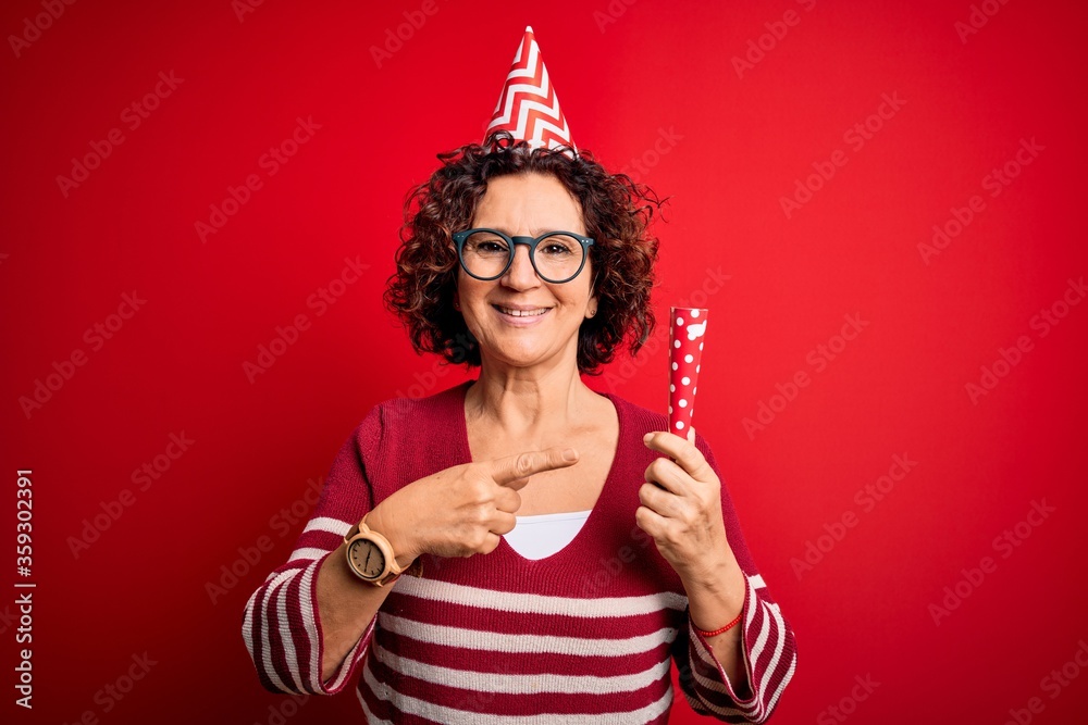 Middle age curly hair woman wearing birthday funny hat holding party trumpet on celebration very happy pointing with hand and finger Middle age curly hair woman wearing birthday funny hat holding party trumpet on celebration very happy pointing with hand and finger