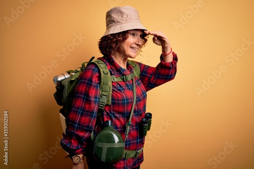 Middle age curly hair hiker woman hiking wearing backpack and water canteen using binoculars very happy and smiling looking far away with hand over head. Searching concept.