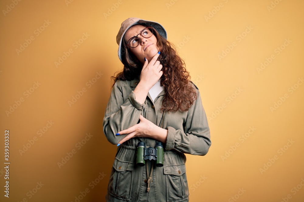 Young beautiful tourist woman on vacation wearing explorer hat and binoculars with hand on chin thinking about question, pensive expression. Smiling with thoughtful face. Doubt concept. Young beautiful tourist woman on vacation wearing explorer hat and binoculars with hand on chin thinking about question, pensive expression. Smiling with thoughtful face. Doubt concept.