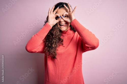 Young beautiful woman with curly hair wearing turtleneck sweater over pink background doing ok gesture like binoculars sticking tongue out, eyes looking through fingers. Crazy expression.