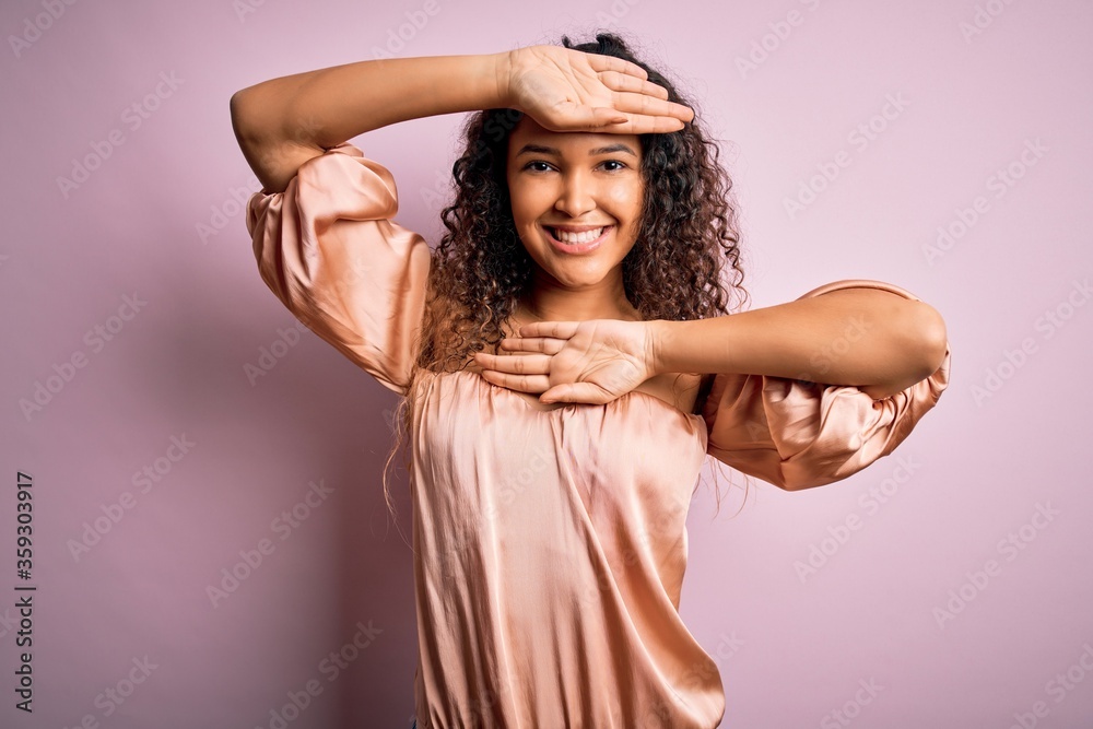 Naklejka premium Young beautiful woman with curly hair wearing casual t-shirt standing over pink background Smiling cheerful playing peek a boo with hands showing face. Surprised and exited