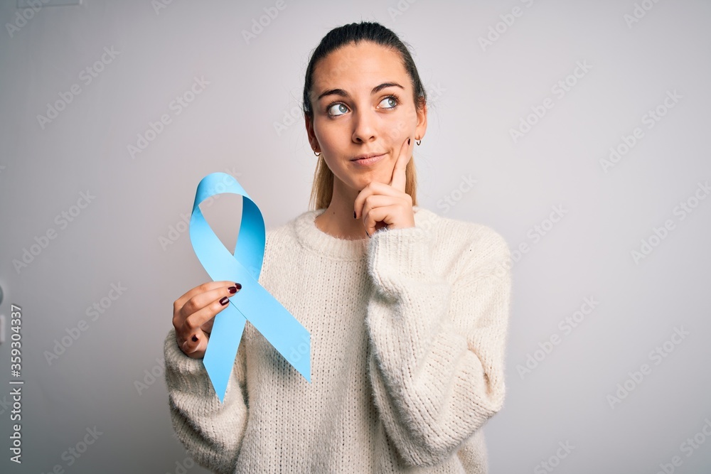 Young beautiful blonde woman holding blue cancer ribbon over isolated white background serious face thinking about question, very confused idea Young beautiful blonde woman holding blue cancer ribbon over isolated white background serious face thinking about question, very confused idea