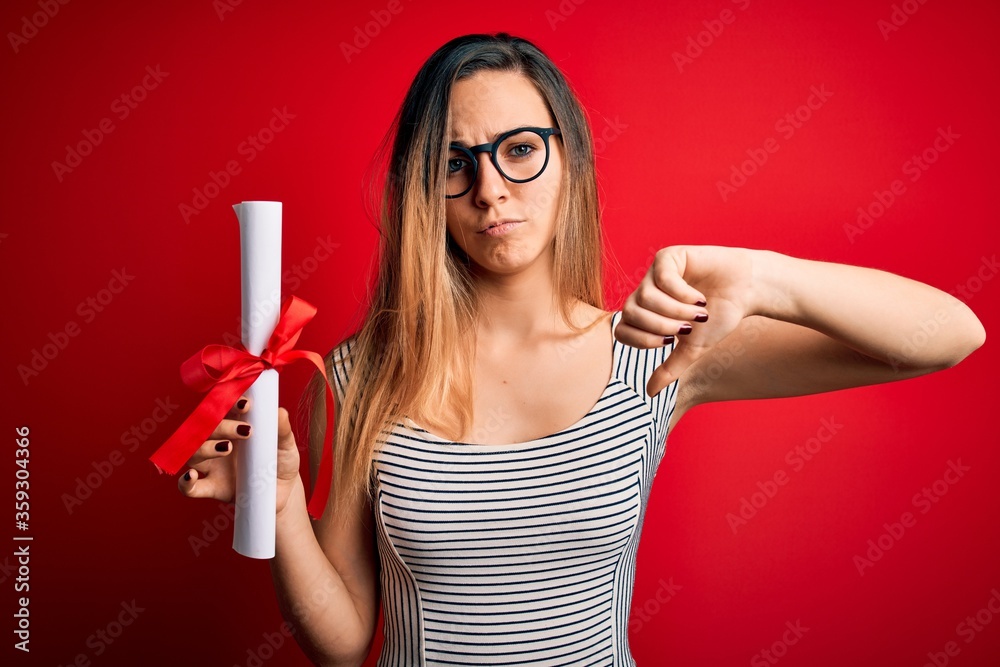 Young blonde woman with blue eyes wearing glasses holding graduated degree diploma with angry face, negative sign showing dislike with thumbs down, rejection concept Young blonde woman with blue eyes wearing glasses holding graduated degree diploma with angry face, negative sign showing dislike with thumbs down, rejection concept