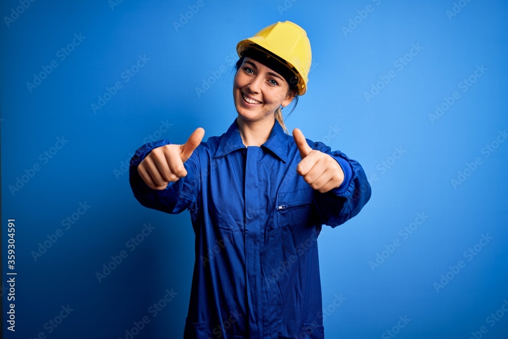 Young beautiful worker woman with blue eyes wearing security helmet and uniform approving doing positive gesture with hand, thumbs up smiling and happy for success. Winner gesture.
