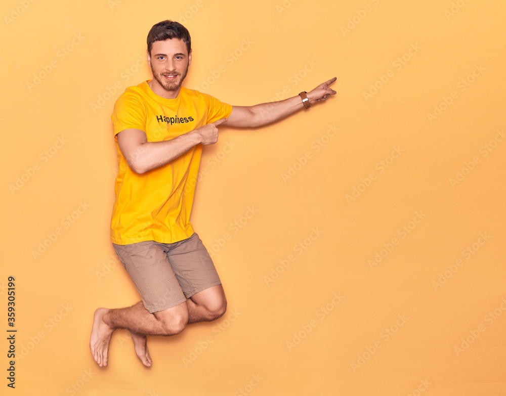 Young handsome hispanic man wearing t shirt with happiness word message smiling happy. Jumping with smile on face over isolated yellow background