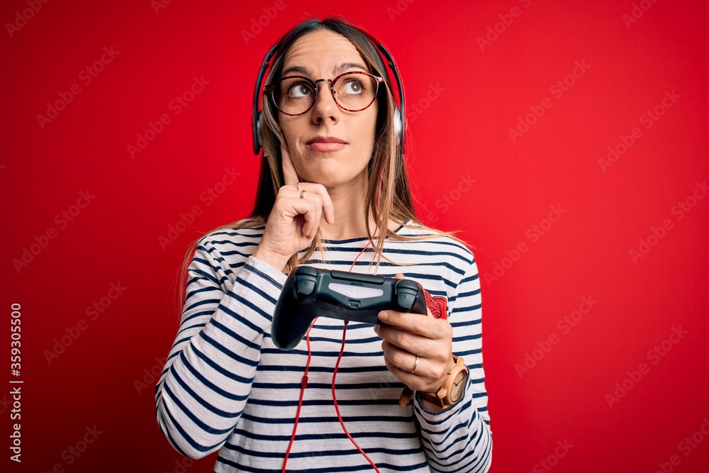 Young blonde gamer woman using gamepad playing video games over red background serious face thinking about question, very confused idea Young blonde gamer woman using gamepad playing video games over red background serious face thinking about question, very confused idea
