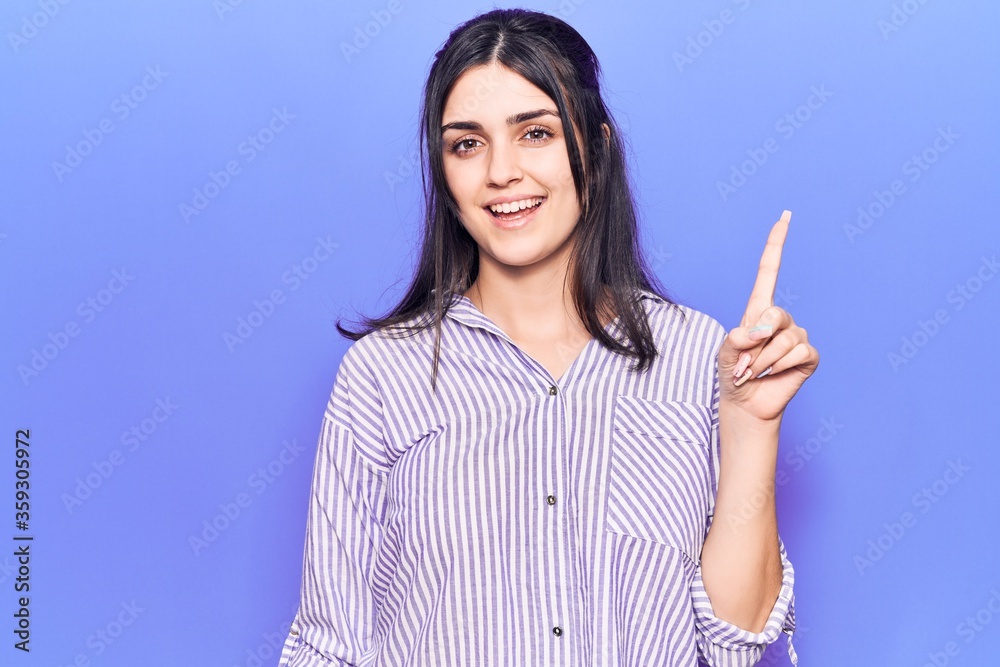Young beautiful girl wearing casual striped shirt smiling with an idea or question pointing finger up with happy face, number one Young beautiful girl wearing casual striped shirt smiling with an idea or question pointing finger up with happy face, number one