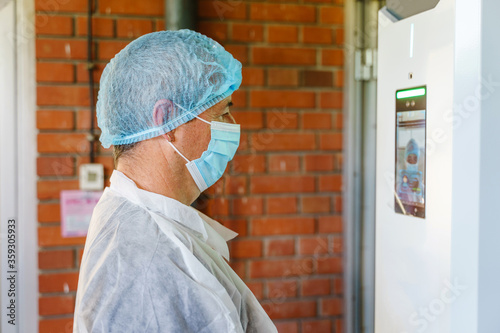 Side view of caucasian man doctor scientist wearing protective gear standing in front of electronic face recognition terminal with digital temperature control camera checking in before going to work photo