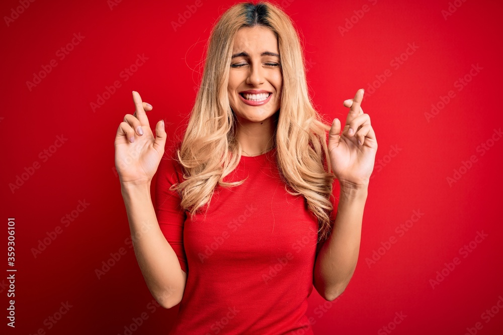 Young beautiful blonde woman wearing casual t-shirt standing over isolated red background gesturing finger crossed smiling with hope and eyes closed. Luck and superstitious concept. Young beautiful blonde woman wearing casual t-shirt standing over isolated red background gesturing finger crossed smiling with hope and eyes closed. Luck and superstitious concept.
