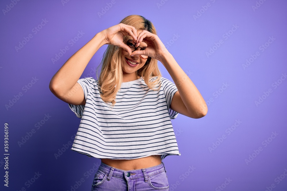 Young beautiful blonde woman wearing striped t-shirt and glasses over purple background Doing heart shape with hand and fingers smiling looking through sign Young beautiful blonde woman wearing striped t-shirt and glasses over purple background Doing heart shape with hand and fingers smiling looking through sign