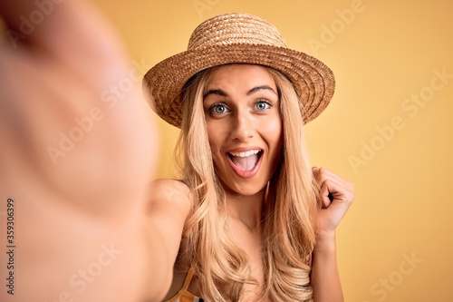 Young beautiful blonde woman on vacation wearing bikini and hat making selfie by camera screaming proud and celebrating victory and success very excited, cheering emotion
