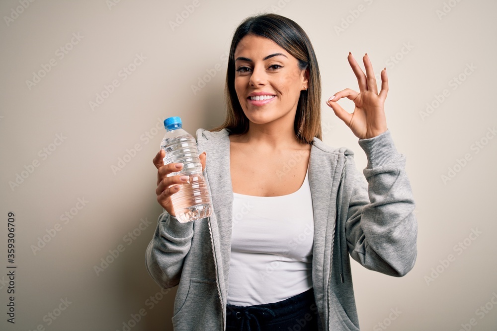 Young beautiful brunette sporty woman drinking bottle of water over isolated white background doing ok sign with fingers, excellent symbol Young beautiful brunette sporty woman drinking bottle of water over isolated white background doing ok sign with fingers, excellent symbol