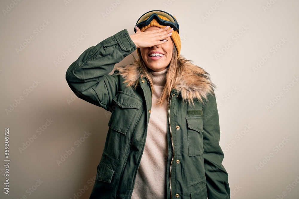 Young brunette skier woman wearing snow clothes and ski goggles over white background smiling and laughing with hand on face covering eyes for surprise. Blind concept.