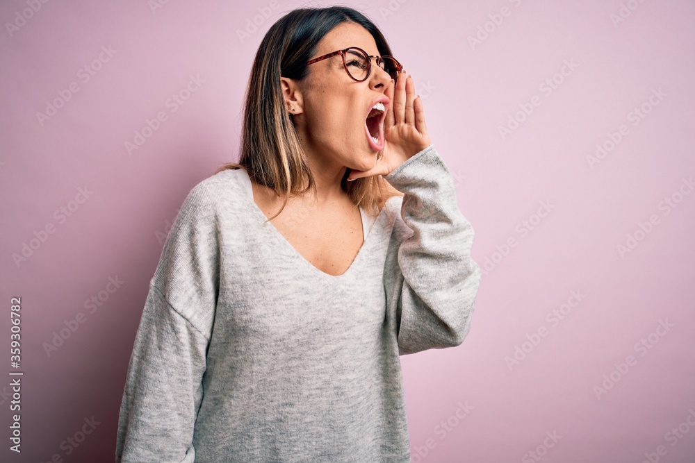 Young beautiful brunette woman wearing casual sweater and glasses over pink background shouting and screaming loud to side with hand on mouth. Communication concept.