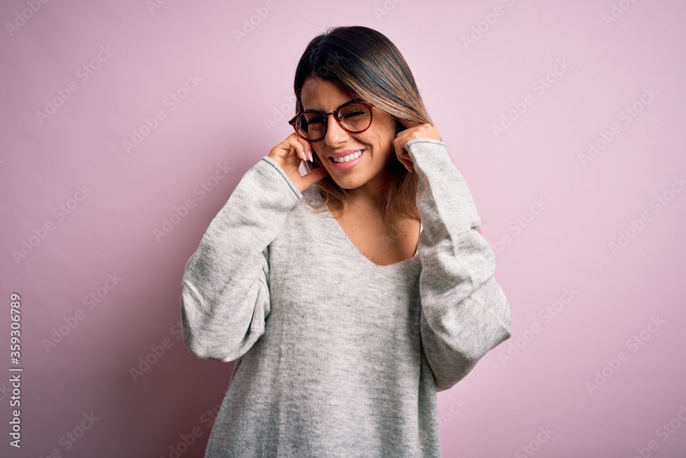 Young beautiful brunette woman wearing casual sweater and glasses over pink background covering ears with fingers with annoyed expression for the noise of loud music. Deaf concept. Young beautiful brunette woman wearing casual sweater and glasses over pink background covering ears with fingers with annoyed expression for the noise of loud music. Deaf concept.