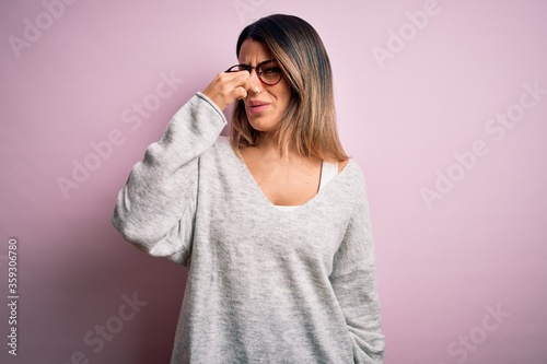 Young beautiful brunette woman wearing casual sweater and glasses over pink background smelling something stinky and disgusting, intolerable smell, holding breath with fingers on nose. Bad smell