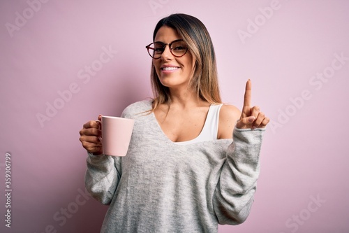 Young beautiful brunette woman wearing glasses drinking pink mug of coffee surprised with an idea or question pointing finger with happy face, number one