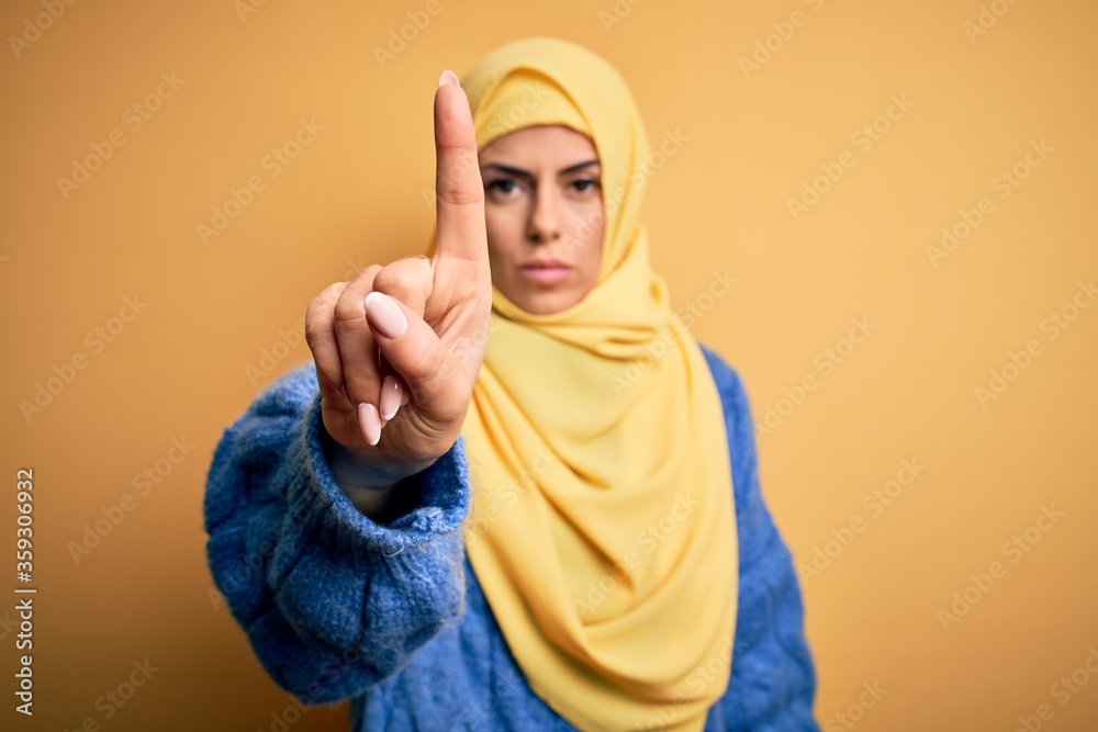 Young beautiful brunette muslim woman wearing arab hijab over isolated yellow background Pointing with finger up and angry expression, showing no gesture Young beautiful brunette muslim woman wearing arab hijab over isolated yellow background Pointing with finger up and angry expression, showing no gesture