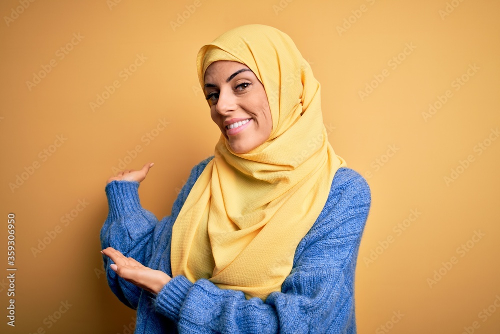 Obraz premium Young beautiful brunette muslim woman wearing arab hijab over isolated yellow background Inviting to enter smiling natural with open hand