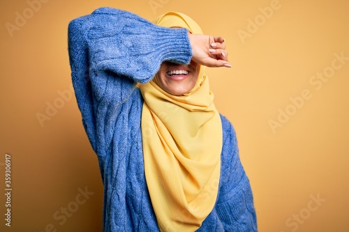 Young beautiful brunette muslim woman wearing arab hijab over isolated yellow background covering eyes with arm smiling cheerful and funny. Blind concept. © Krakenimages.com