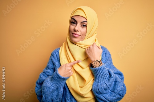 Young beautiful brunette muslim woman wearing arab hijab over isolated yellow background In hurry pointing to watch time, impatience, looking at the camera with relaxed expression
