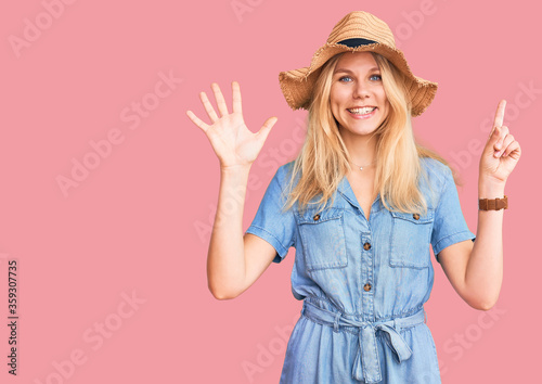 Young beautiful blonde woman wearing summer hat and dress showing and pointing up with fingers number six while smiling confident and happy.
