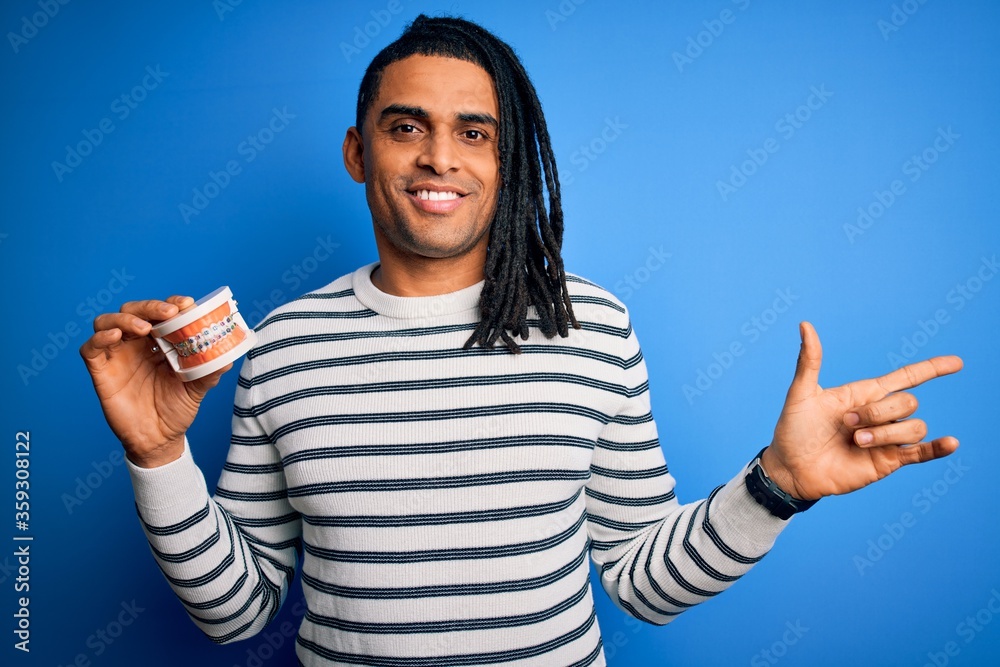 Young african american afro man holding plastic teeth with dental braces very happy pointing with hand and finger to the side Young african american afro man holding plastic teeth with dental braces very happy pointing with hand and finger to the side
