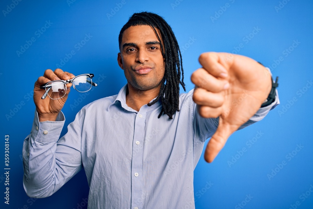 Fototapeta premium Young african american afro man with dreadlocks holding glasses over blue background with angry face, negative sign showing dislike with thumbs down, rejection concept