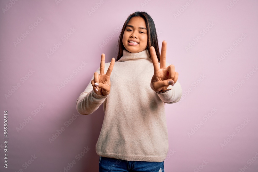 Young beautiful asian girl wearing casual turtleneck sweater over isolated pink background smiling looking to the camera showing fingers doing victory sign. Number two. Young beautiful asian girl wearing casual turtleneck sweater over isolated pink background smiling looking to the camera showing fingers doing victory sign. Number two.
