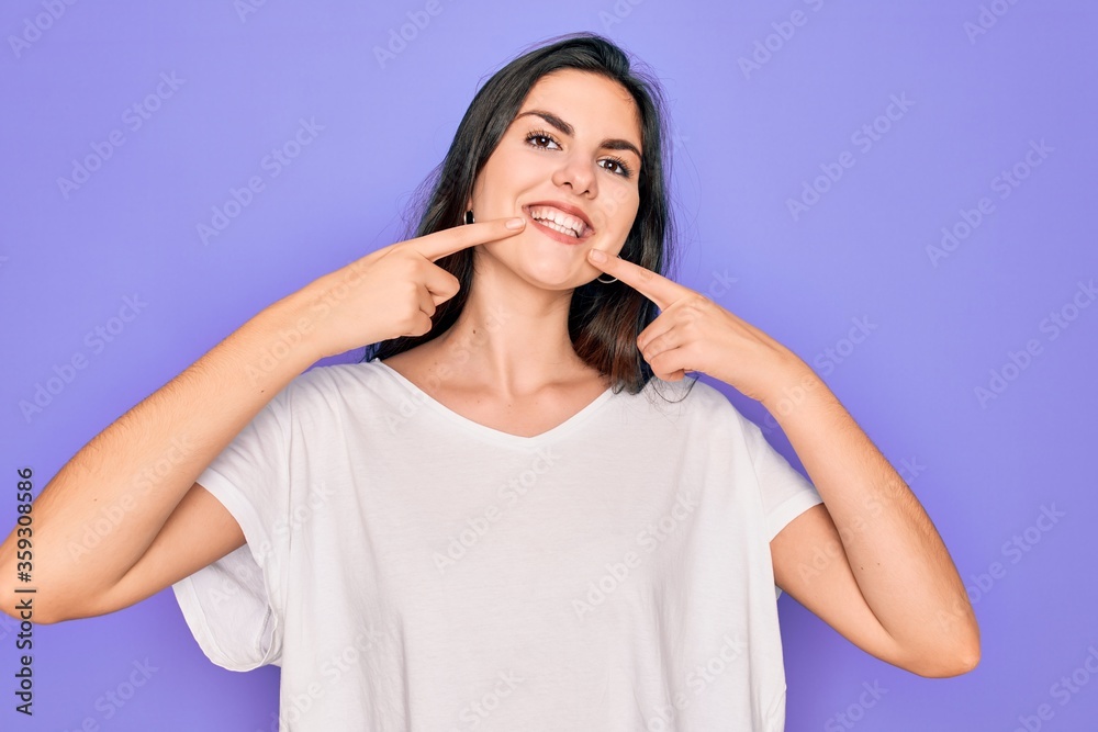 Young beautiful brunette woman wearing casual white t-shirt over purple background smiling cheerful showing and pointing with fingers teeth and mouth. Dental health concept. Young beautiful brunette woman wearing casual white t-shirt over purple background smiling cheerful showing and pointing with fingers teeth and mouth. Dental health concept.
