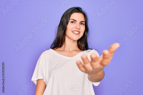 Young beautiful brunette woman wearing casual white t-shirt over purple background smiling cheerful offering palm hand giving assistance and acceptance.