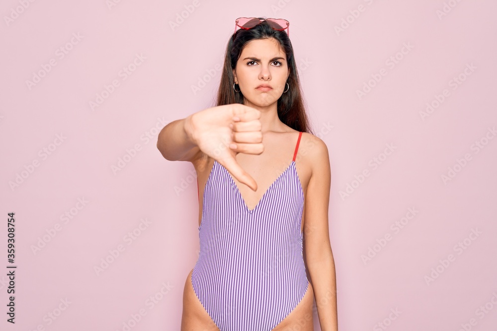 Obraz premium Young beautiful fashion girl wearing swimwear swimsuit and sunglasses over pink background looking unhappy and angry showing rejection and negative with thumbs down gesture. Bad expression.