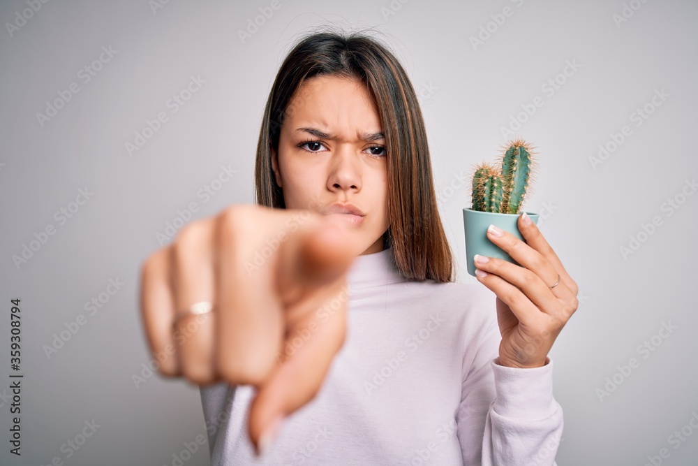 Young beautiful brunette girl holding small cactus plant pot over isolated white background pointing with finger to the camera and to you, hand sign, positive and confident gesture from the front Young beautiful brunette girl holding small cactus plant pot over isolated white background pointing with finger to the camera and to you, hand sign, positive and confident gesture from the front
