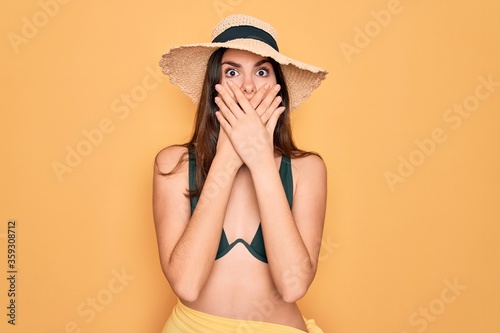 Young beautiful girl wearing swimwear bikini and summer sun hat over yellow background shocked covering mouth with hands for mistake. Secret concept.