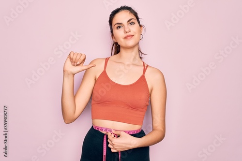 Young beautiful fitness woman wearing sport excersie clothes using measuring tape with surprise face pointing finger to himself photo