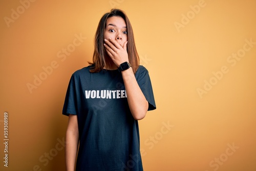 Young beautiful brunette girl doing volunteering wearing t-shirt with volunteer message word shocked covering mouth with hands for mistake. Secret concept.