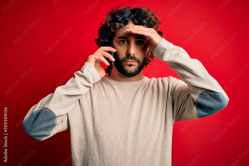 Handsome man with beard having conversation talking on the smartphone over red background stressed with hand on head, shocked with shame and surprise face, angry and frustrated. Fear and upset.